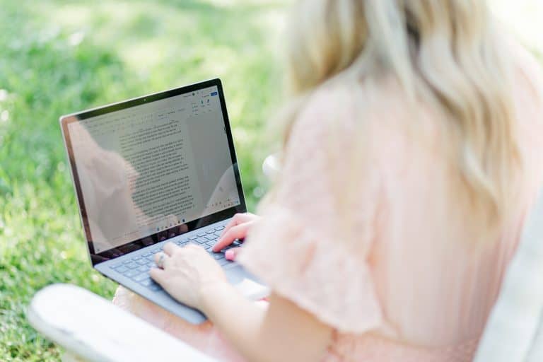 A view of a computer screen on the lap of Brettnay Brazzell. She is sitting, facing away from the camera and typing. She is in a white rocking chair with grass showing in the background