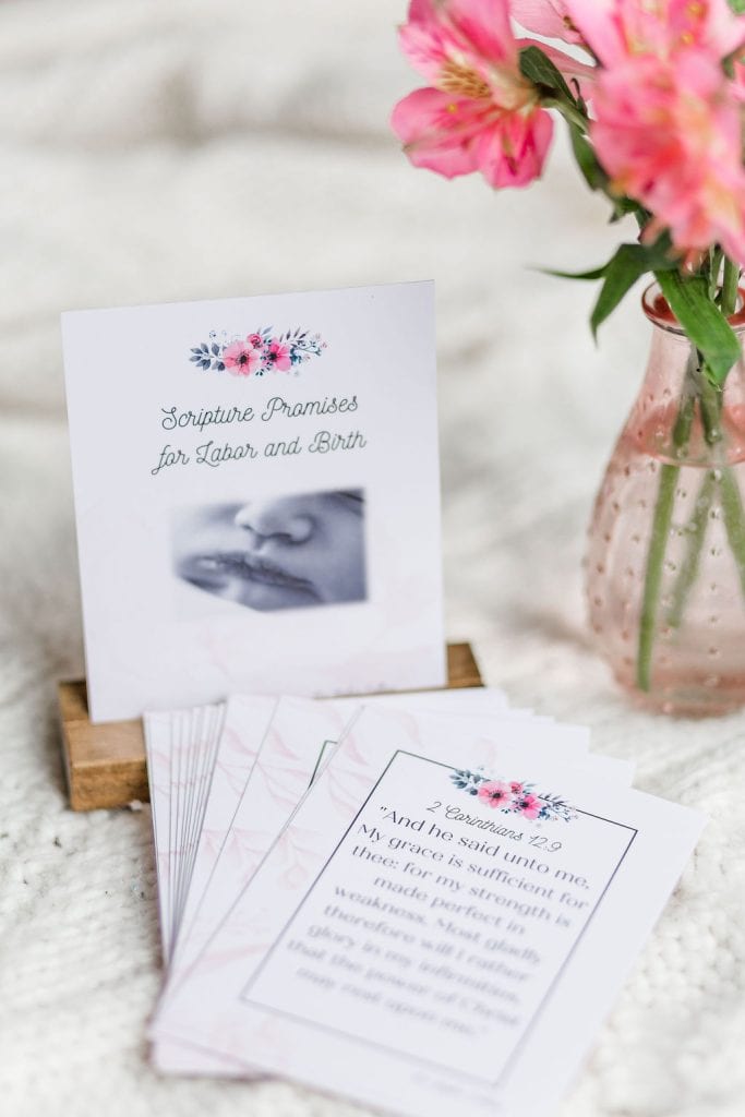 labor and delivery cards. It depicts the front card in a wooden stand with other cards flayed out in front of it. They are laying on a white blanket with a small vase of pink flowers beside them.