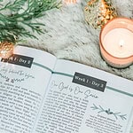 Great Hymns for Growing Hearts - Christmas Edition. The book is open to display the devotional content page. It is laying on a furry, white rug with a candle and evergreens around it.