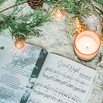 Great Hymns for Growing Hearts - Christmas Edition. The book is open to show the music to "Silent Night." It is laying on a furry, white rug with a candle and evergreens around it.