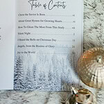 Great Hymns for Growing Hearts - Christmas Edition. The book is open to the table of contents. It is laying on a furry, white rug with a candle and evergreens around it.