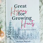 Great Hymns for Growing Hearts - Christmas Edition. The book has a snowy forest background. It is laying on a furry, white rug with a candle and evergreens around it.