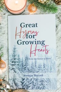 Great Hymns for Growing Hearts - Christmas Edition. The book has a snowy forest background. It is laying on a furry, white rug with a candle and evergreens around it.