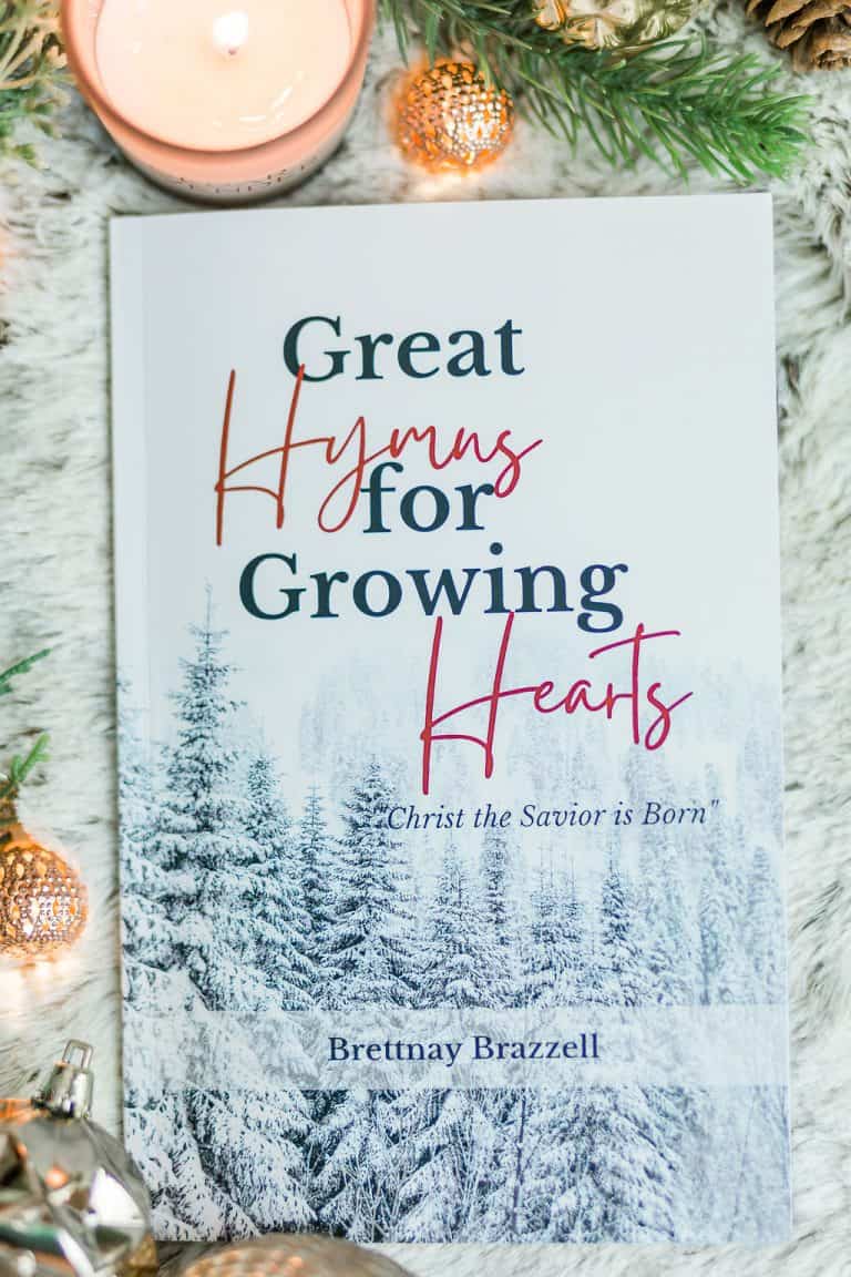 Great Hymns for Growing Hearts - Christmas Edition. The book has a snowy forest background. It is laying on a furry, white rug with a candle and evergreens around it.