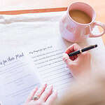 The Head to Toe Prayer Journal laying open on a table. A woman's hands are shown writing in it. There is a plant, pens, and a coffee next to the book.