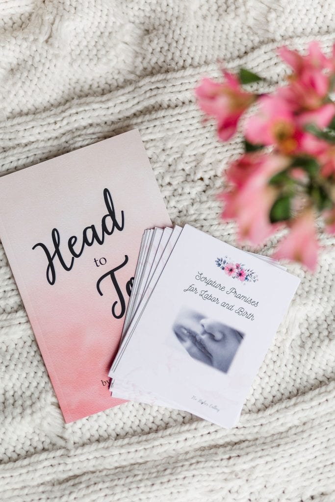 The "Head-to-Toe" prayer journal and the "Scripture Promises Labor and Birth" cards. The cards are laying on top and to the side of the book. They are laying on a white blanket with flowers in the foreground.