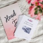 The "Head-to-Toe" prayer journal and the "Scripture Promises Labor and Birth" cards. The cards are laying on top and to the side of the book. They are laying on a white blanket with flowers in the foreground.