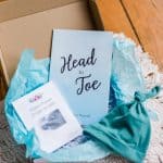 New Mama Gift Box in blue including a Head-to-Toe Prayer Journal, the "Scripture Promises Labor and Birth Cards" and a baby hat all in a brown box sitting on a white mat on a wooden floor.