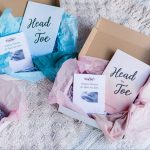 Two gift boxes, one pink and one blue, on a white blanket. They include the Head-to-Toe Prayer Journals, the Scripture Promise Labor and Birth Cards, and a baby hat.