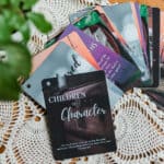 The Children of Character Cards with the ring removed and spread out on a table. The cards are several colors and have various background images. The table has the cards, a plant, and a candle on it.