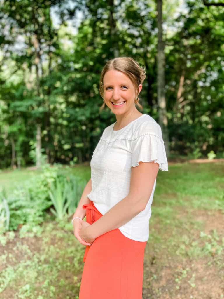 Sarah Boots, wearing a white top and peach skirt, is standing with hands in lap in front of a green field. There are trees lining the backdrop.