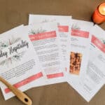 The Holiday Hospitality Guide laying out on a brown floor. There are a few images and parts of recipes that can be seen. There is a wooden spoon and a candle next to it.
