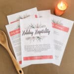 The Holiday Hospitality Guide laying out on a brown background. Next to it are a candle which is lit and a wooden spoon.