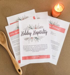 The Holiday Hospitality Guide laying out on a brown background. Next to it are a candle which is lit and a wooden spoon.
