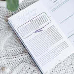 Great Hymns for Growing Hearts book interior image. It shows the hymn's story on one side and a section called "Heart Application" on the right. The book sits on a table with a plant near it.