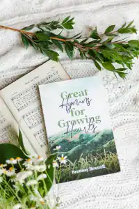 Great Hymns for Growing Hearts book laying on a table with flowers around it. On the table is a hymn book laying open as well.