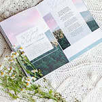 Great Hymns for Growing Hearts book laying open on a table beside a coffee and a bunch of flowers. Interior page shows a hymn title on the left and the words on the right. There is a image of woods and the setting sun in the background.