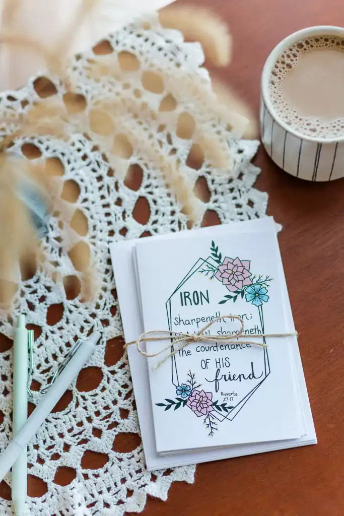 A stack of Friendship Cards wrapped in twin with a bow. It is sitting on a hardwood table covered in lace along side a few pens.