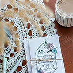 A stack of Friendship Cards wrapped in twin with a bow. It is sitting on a hardwood table covered in lace along side a few pens.