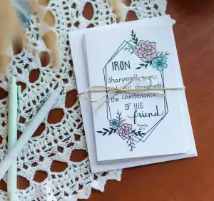 A stack of Friendship Cards wrapped in twin with a bow. It is sitting on a hardwood table covered in lace along side a few pens.