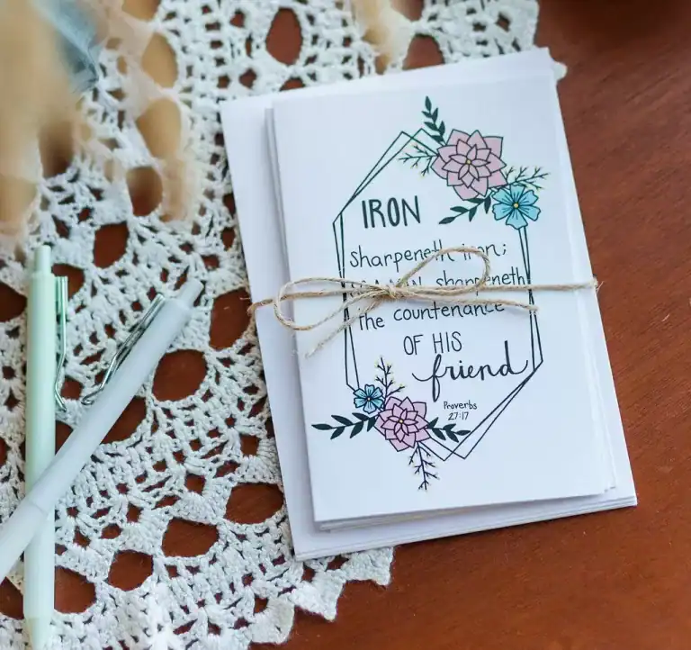 A stack of Friendship Cards wrapped in twin with a bow. It is sitting on a hardwood table covered in lace along side a few pens.
