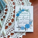 A Friendship card laying on a wooden table with a lace cover. The card reads "Ointment and perfume rejoice the heart: so doth the sweetness of a man's friend by hearty counsel." Proverbs 27:9