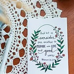 A Friendship Card laying on a wooden table with a lace covering. The card reads "And let us consider one another to provoke unto love and good works:" Hebrews 10:25. The text is wrapped in flowers and leaves