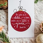 A single card laying on fir and surrounded by greenery. It has a red Christmas bulb and says "For unto us a child is born, unto us a Son is given" Isaiah 9:6
