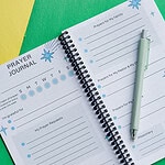 The Teach Me To Pray Prayer Journal laying on 2 pieces of construction paper, one yellow and one green. It is open with a pen laying on it to show the prayer requests and prompts area. There are colored pencils next to it.