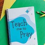 The "Teach Me to Pray Prayer Journal for Kids" laying on two sheets of yellow and green construction paper with colored pencils next to it.