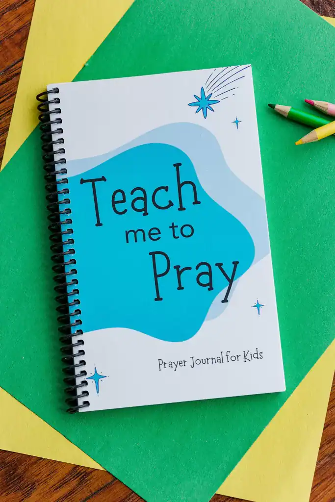The "Teach Me to Pray Prayer Journal for Kids" laying on two sheets of yellow and green construction paper with colored pencils next to it.