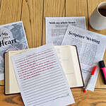 The Psalm 119 writing plan beneath a bible and a written on notepage along side a red pen. They are all sitting on a wooden table.