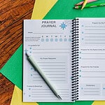 The Teach Me To Pray Prayer Journal laid open to show the prayer request and prompt areas. It is on top of colored construction paper on a wooden table. There are colored pencils beside it.